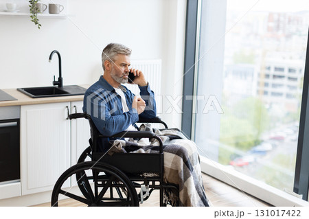 Senior Caucasian man sitting in wheelchair by kitchen indoors using phone near window. Scene depicts everyday lifestyle, accessibility, comfort, connection, communication, independence, and inclusion Senior Caucasian man sitting in wheelchair by kitchen indoors using phone near window. Scene depicts everyday lifestyle, accessibility, comfort, connection, communication, independence, and inclusion 131017422