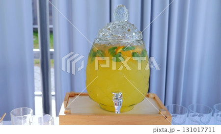 Glass beverage dispenser with yellow lemonade, orange slices and mint leaves, placed on a wooden crate, perfect for summer parties or gatherings 131017711