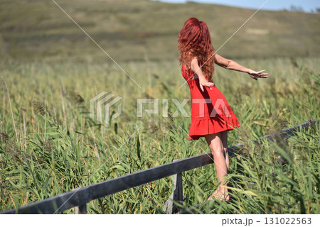 Woman with red hair in red dress walking through scenic natural wetland pathway Woman with red hair in red dress walking through scenic natural wetland pathway 131022563