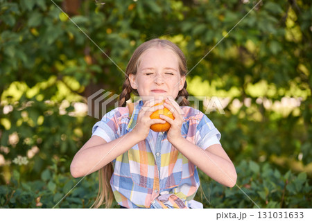 Caucasian young girl enjoying a fresh orange outdoors in vibrant sunlit garden 131031633