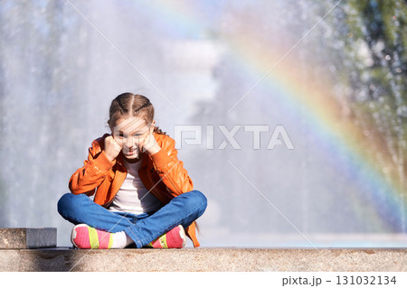 Smiling caucasian female child sitting by fountain with rainbow in bright Smiling caucasian female child sitting by fountain with rainbow in bright 131032134