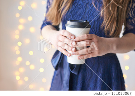 Young caucasian female in blue dress holding coffee cup with festive lights Young caucasian female in blue dress holding coffee cup with festive lights 131033067