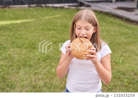 Caucasian young girl eagerly biting into a large round bread in a garden setting Caucasian young girl eagerly biting into a large round bread in a garden setting 131033259