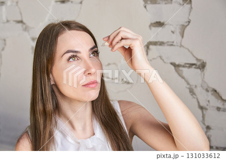 Young caucasian female applying eye drops in front of cracked wall background 131033612
