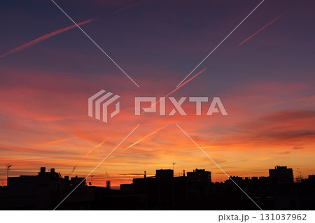 A vibrant sunset with shades of orange, pink, and purple fills the sky. Silhouettes of buildings and contrails are visible against the colorful backdrop. 131037962