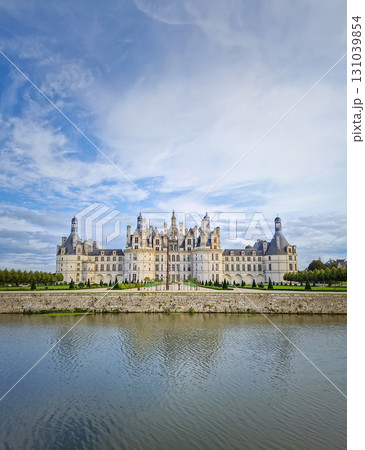The symmetrical Chateau de Chambord is reflected in the still water, highlighting its magnificent Renaissance architecture with numerous turrets. The majestic castle stands beneath a beautiful sky 131039854