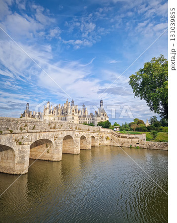 A picturesque stone bridge spans the still water leading to the magnificent Chateau de Chambord. The iconic French Renaissance castle stands under a beautiful sky with soft clouds 131039855