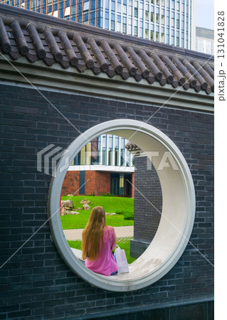 young woman sits in a round window in the fence of a Chinese garden young woman sits in a round window in the fence of a Chinese garden 131041828