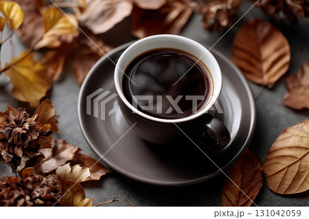 Warm cup of black coffee with autumn leaves and cozy atmosphere, dark ceramic cup on saucer surrounded by dried brown leaves and seasonal pinecones 131042059