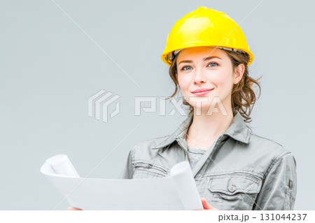 Female construction worker holding blueprints while wearing a yellow hard hat in a bright environment 131044237
