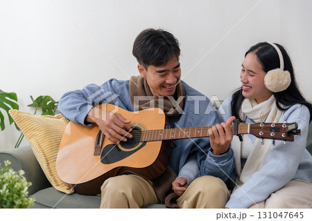Winter day finds happy couple at home, young people enjoying relaxing indoor comfort, man playing acoustic guitar for smiling woman, warm sweaters cozy scarf comfort chilly winter day. 131047645