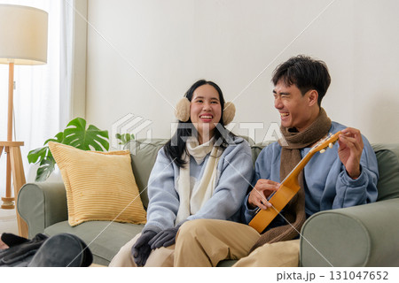 Winter day brings happy couple, family members relaxing indoor home with warm scarves and earmuffs, playing ukulele, sharing laughter, comfortable moment creating cozy atmosphere cold winter season. 131047652