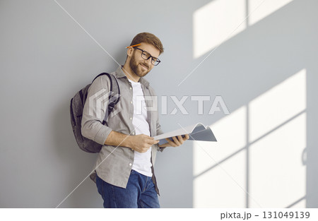 Portrait of happy male student with textbook and backpack standing by grey wall background Portrait of happy male student with textbook and backpack standing by grey wall background 131049139