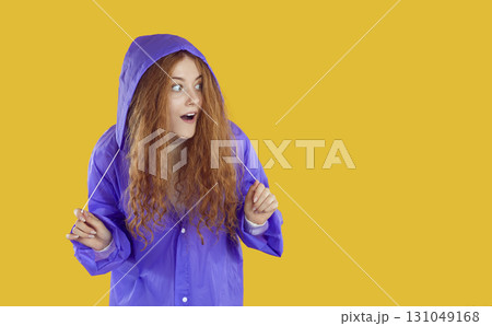 Amazed redhead girl in purple coat looks out from under the hood on copy space on yellow background. 131049168
