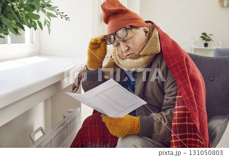 Senior man in warm clothes sitting by cold radiator and looking at expensive heating bill Senior man in warm clothes sitting by cold radiator and looking at expensive heating bill 131050803