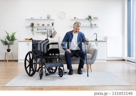 Mature adult male working towards mobility recovery with cane beside wheelchair in living room Mature adult male working towards mobility recovery with cane beside wheelchair in living room 131053140