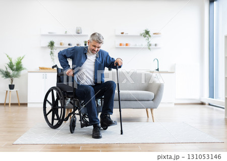 Disabled adult man practicing walking indoors using cane for support near wheelchair. Home environment emphasizes independence, determination, and adaptive rehabilitation. 131053146