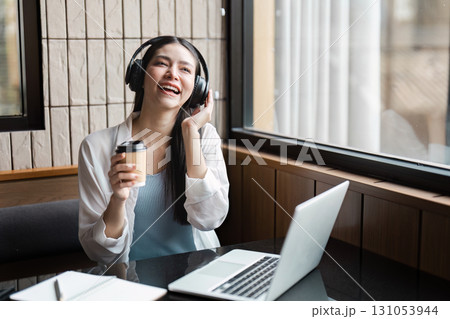 Young professional woman enjoying music while working on a laptop in a modern cafe, promoting productivity and relaxation. Young professional woman enjoying music while working on a laptop in a modern cafe, promoting productivity and relaxation. 131053944