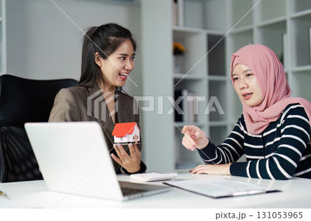 Real estate agent and client discussing property options with a model house in a stylish office environment. 131053965