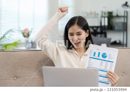 Excited young woman celebrating success while reviewing financial reports at home. 131053994
