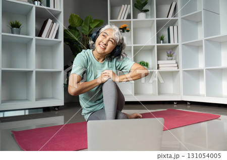 Senior woman enjoying wellness activities at home, listening to music with headphones while sitting on yoga mat. 131054005