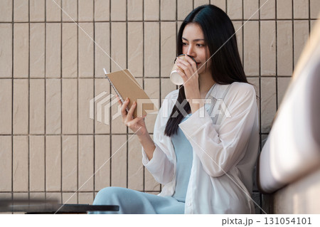 Young Asian woman enjoying coffee while reading a notebook in a cozy cafe setting. 131054101