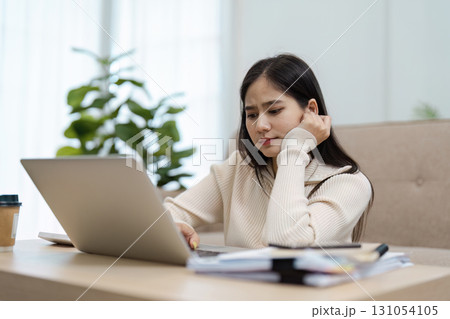 Young Asian woman looking concerned while working on a laptop at home, reflecting on challenges. 131054105