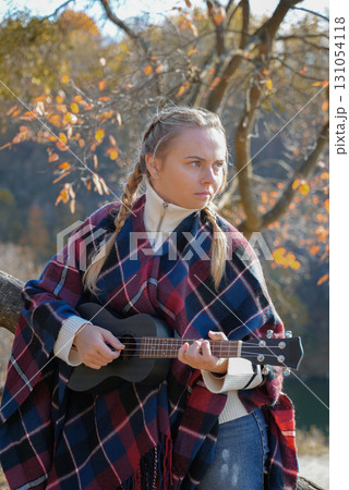 Portrait of smiling caucasian girl plays ukulele in autumnal park. Young woman plays guitar musical instrument outside in nature fall time. Audio music healing Portrait of smiling caucasian girl plays ukulele in autumnal park. Young woman plays guitar musical instrument outside in nature fall time. Audio music healing 131054118