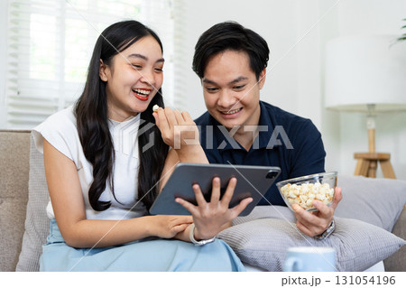 Couple enjoying a movie together on a tablet, sharing laughter and popcorn in a cozy living room setting. Couple enjoying a movie together on a tablet, sharing laughter and popcorn in a cozy living room setting. 131054196