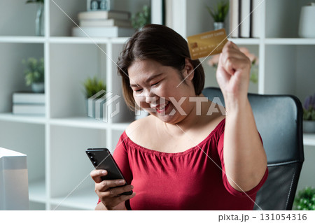 Excited woman showing her credit card while shopping online in a cozy workspace. Excited woman showing her credit card while shopping online in a cozy workspace. 131054316