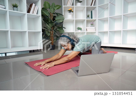 Senior woman performing yoga poses with headphones while following an online session at home. Senior woman performing yoga poses with headphones while following an online session at home. 131054451