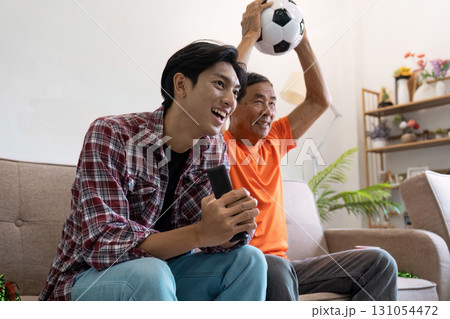Joyful Asian father and son celebrating a soccer match at home, showcasing family excitement and sports bonding. 131054472