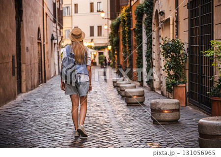 Teenage girl with long hair in a straw hat and backpack walking along a charming cobblestone street in Rome at sunset 131056965
