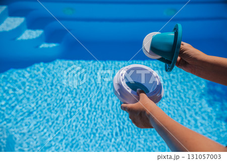 Person placing chlorine tablet into floating dispenser while standing near bright blue swimming pool steps for effective water disinfection 131057003
