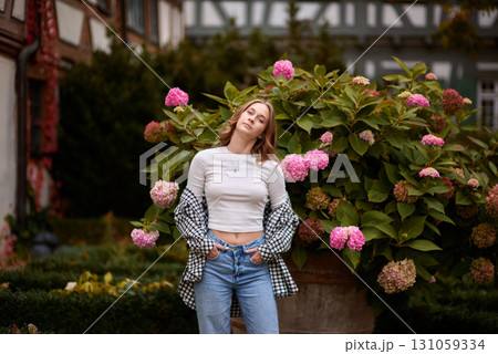 Autumn Fashion Portrait: Stylish Woman Amidst Blooming Hydrangeas at Traditional German Half Timbered House. Cozy Fall Outdoor Scene Showcasing Casual Outfit and Warm Colors in Picturesque Rural 131059334