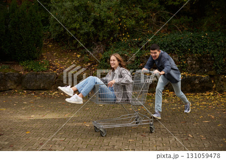 Happy young couple having fun with shopping cart outdoors in autumn park, wearing stylish casual outfits with denim and flannel, laughing and enjoying joyful moment, surrounded by autumn foliage and Happy young couple having fun with shopping cart outdoors in autumn park, wearing stylish casual outfits with denim and flannel, laughing and enjoying joyful moment, surrounded by autumn foliage and 131059478