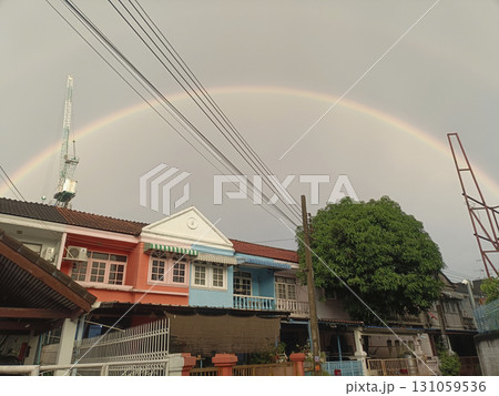 Vibrant rainbow arches across cloudy sky above colorful residential houses with large green tree in foreground, creating peaceful and cheerful atmosphere 131059536