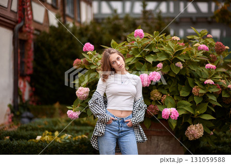 Stylish young woman posing in cozy autumn outfit among blooming hydrangeas in front of traditional half-timbered house in Germany, warm fall colors, outdoor portrait, casual fashion, seasonal 131059588
