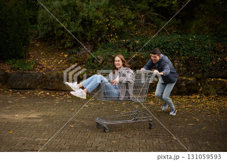 Happy young couple having fun with a shopping cart outdoors in autumn park, wearing stylish casual outfits with denim and flannel, laughing and enjoying the moment. Happy young couple having fun with a shopping cart outdoors in autumn park, wearing stylish casual outfits with denim and flannel, laughing and enjoying the moment. 131059593
