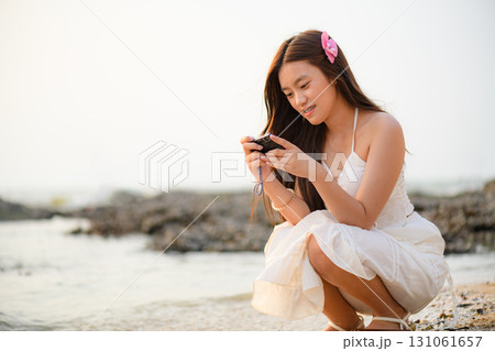 A woman in a white dress is enjoying her vacation on the beach with a retro camera in hand. 131061657