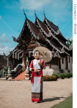 A woman in traditional Thai clothing stands with a parasol in front of an ornate temple under a clear blue sky. 131062311