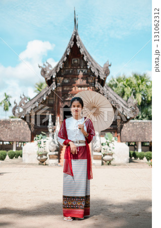 A woman in traditional Thai clothing stands with a parasol in front of an ancient temple, showcasing cultural heritage. 131062312
