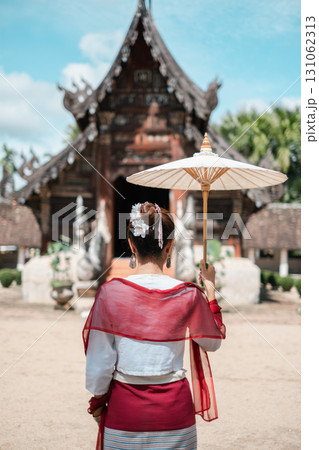 A woman in traditional Thai clothing holds a parasol, standing before an ancient temple under a bright blue sky. 131062313