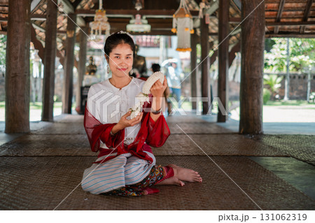 A woman in traditional Thai clothing sits in a wooden pavilion, holding a floral garland, showcasing cultural heritage. 131062319