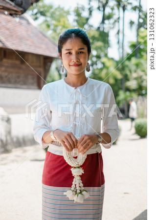 A woman in traditional Thai clothing holds a floral garland, standing outdoors with greenery and traditional architecture in the background. 131062323