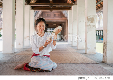 A woman in traditional Thai clothing holds a seashell while sitting in a historic temple corridor, showcasing cultural heritage. 131062326