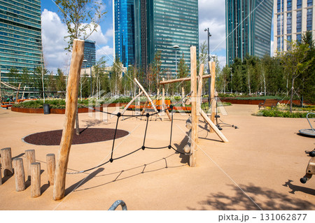 A vibrant playground features wooden structures and swings, set against a backdrop of modern architecture and lush green landscaping during a sunny day. A vibrant playground features wooden structures and swings, set against a backdrop of modern architecture and lush green landscaping during a sunny day. 131062877
