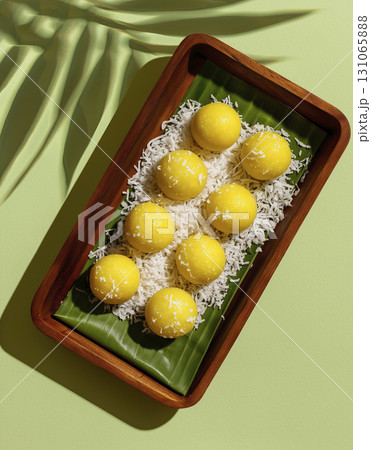Yellow mochi balls with shredded coconut on green banana leaf in wooden tray, bright and inviting presentation, perfect for dessert or snack time 131065888