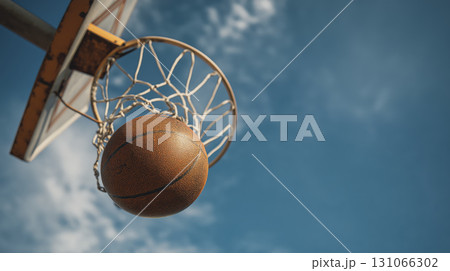 Basketball ball is going through basketball hoop on outdoor court under blue sky with clouds, capturing moment of sports action and excitement 131066302