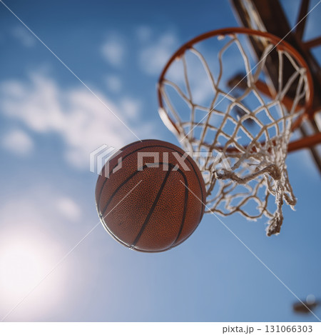 Basketball ball is in mid air near basketball hoop against bright blue sky with clouds, capturing moment of sports action and outdoor activity 131066303